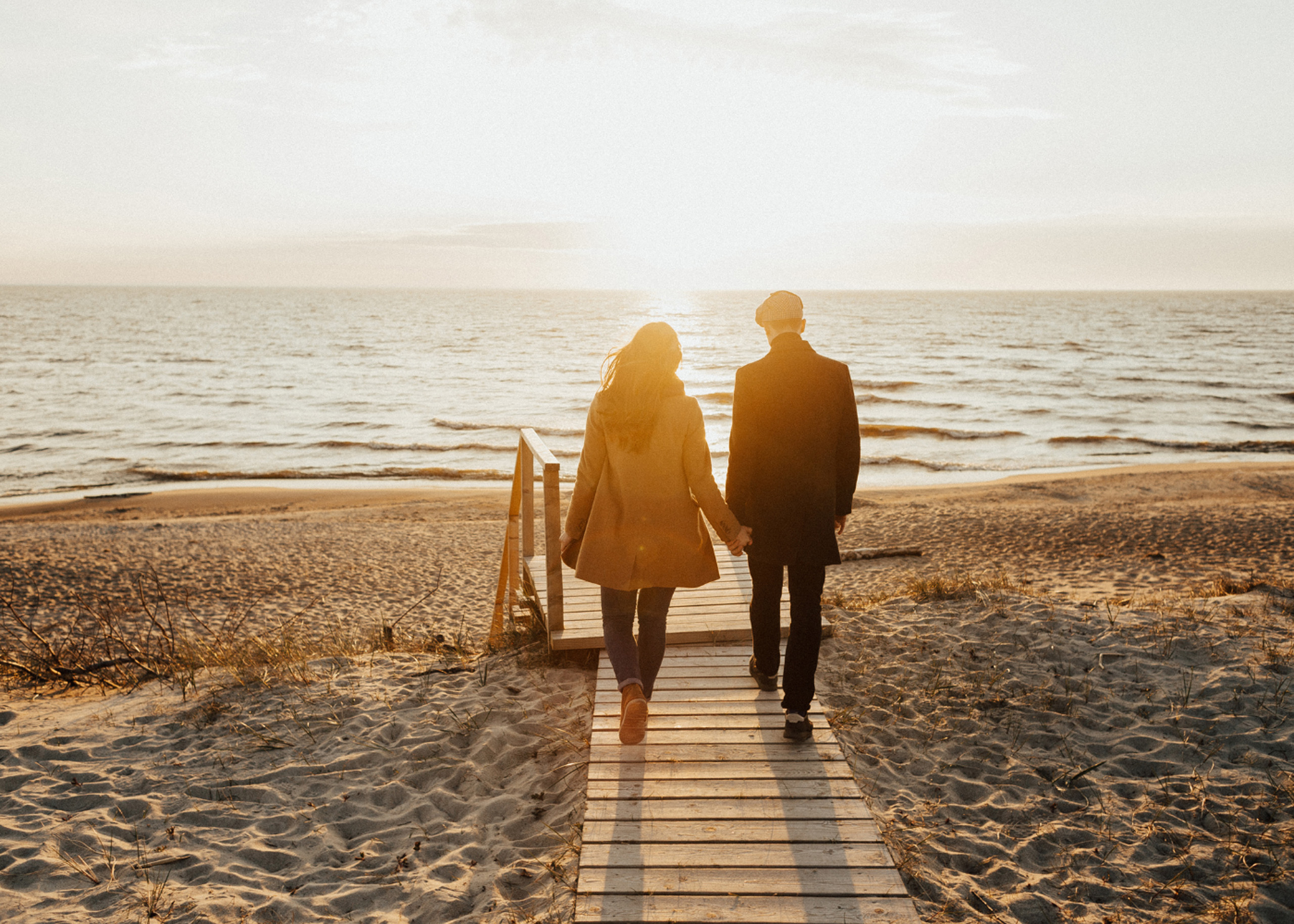Couple holding hands walking toward sunset on boardwalk.