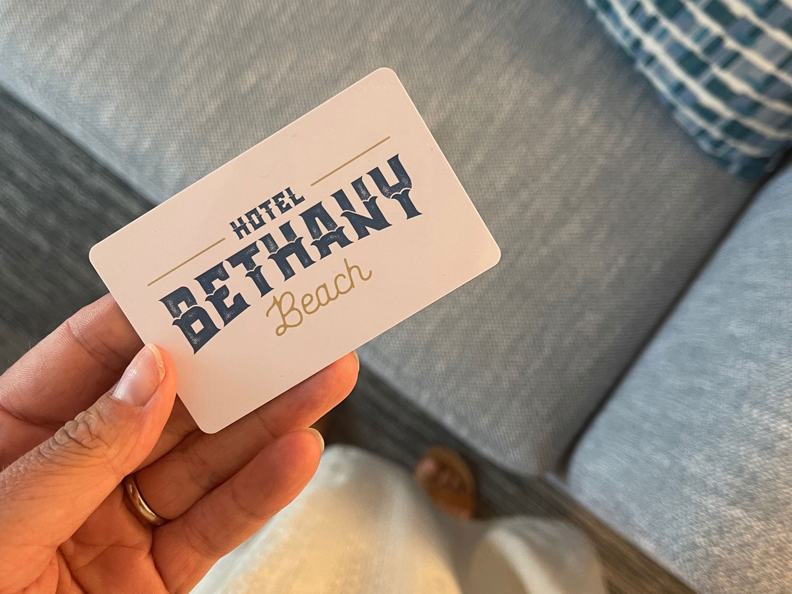 Close-up of a hand holding a Hotel Bethany Beach key card above a blue sofa.