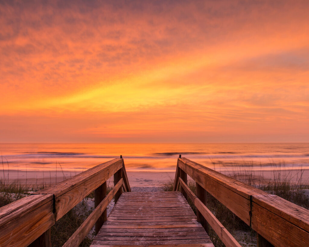 Wooden boardwalk leading to ocean at vibrant sunset.